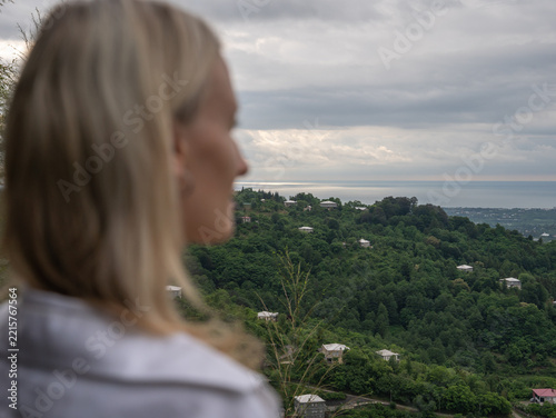 Blonde woman standing on a hill, looking at the panoramic view of the vast green hills reaching the coastal town and the sea under a cloudy sky