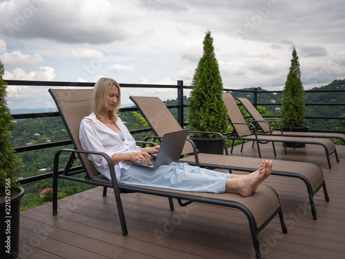 Woman relaxing on a lounge chair, working on her laptop outdoors on a wooden terrace with a scenic view of green mountains and a cloudy sky, embracing the flexible lifestyle of a digital nomad