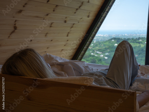 Woman relaxing in bed waking up in a cozy a-frame wooden cabin enjoying a scenic natural view of mountains and the sea, experiencing travel, vacation, and tranquility
