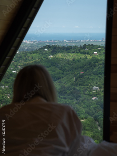 Woman lying on a bed in a modern house, looking out the large window, watching the lush green hills descend to the distant city skyline and the endless blue sea. Vertical photo