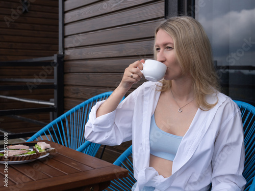 Woman leisurely drinking coffee from a white mug while sitting on a balcony, savoring a calm outdoor breakfast of open sandwiches, embracing relaxation