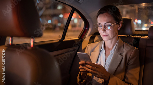 Woman wearing glasses uses a smartphone while sitting in the back seat of a car at night, with blurred city lights in the background.