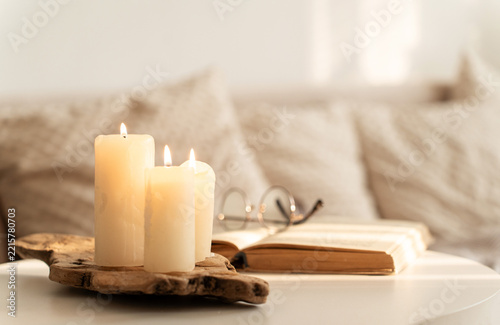 Lit candles on wooden candle holder, an open book, and glasses on white table create warm, cozy home atmosphere. Slow living concept, mindful ritual