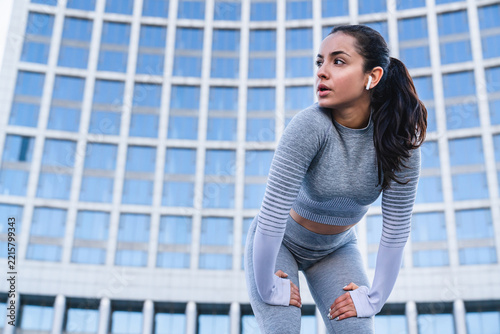 Close-up photo of 20s sportswoman resting after jogging in front of urban background, female athlete recovering outdoors, fitness lifestyle, running, wellness, healthy living active exercise concept