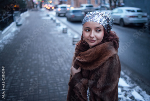 Young woman with red hair wearing a fur coat and sequined headscarf stands on a snowy sidewalk in a city, cars parked along the street in the background