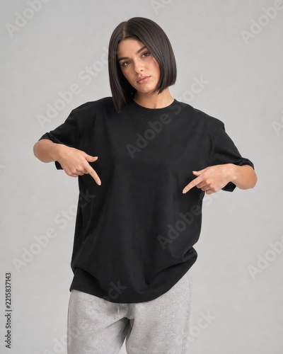 Brunette woman with short dark hair wearing a black t-shirt and gray sweatpants pointing down at her torso.