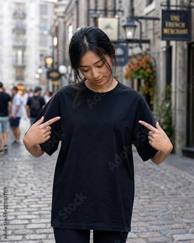 Woman wearing a plain black t-shirt standing on a cobblestone European street pointing down at her chest.