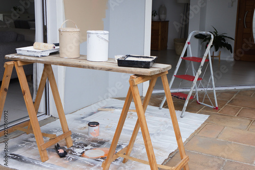 Sawhorses are holding plank bench with beige and white paint cans, red ladder on tiled patio