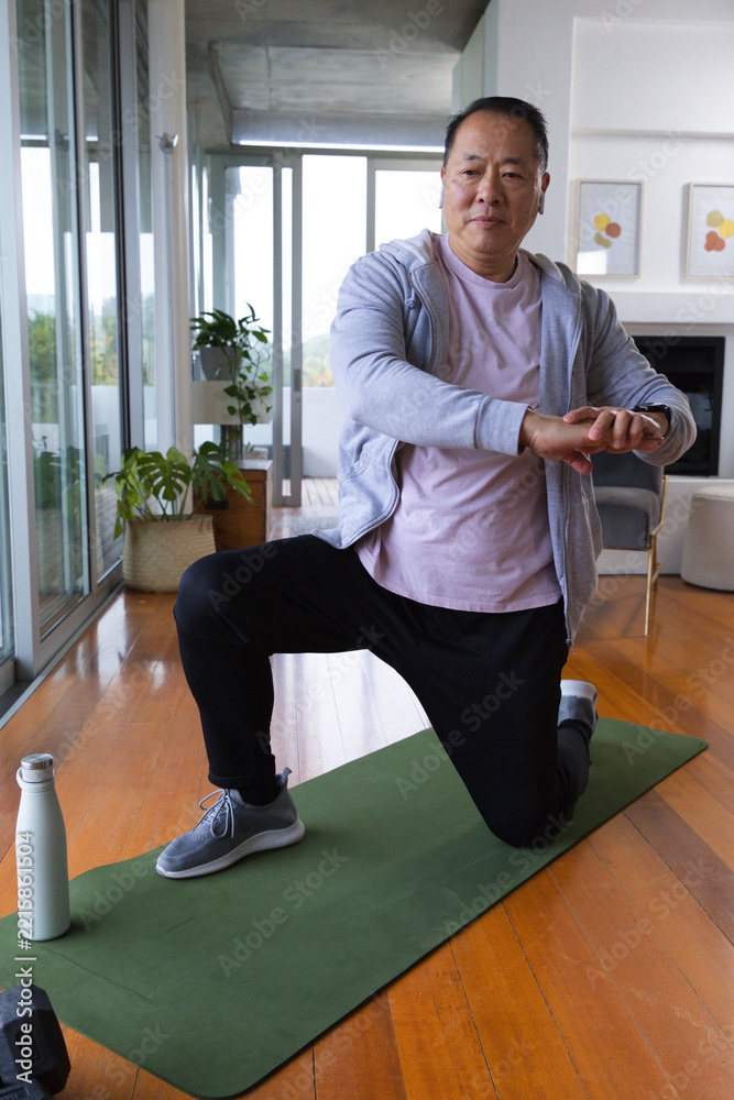 Naklejka premium Senior Asian man in gray hoodie performing kneeling lunge on green mat at home near dumbbell
