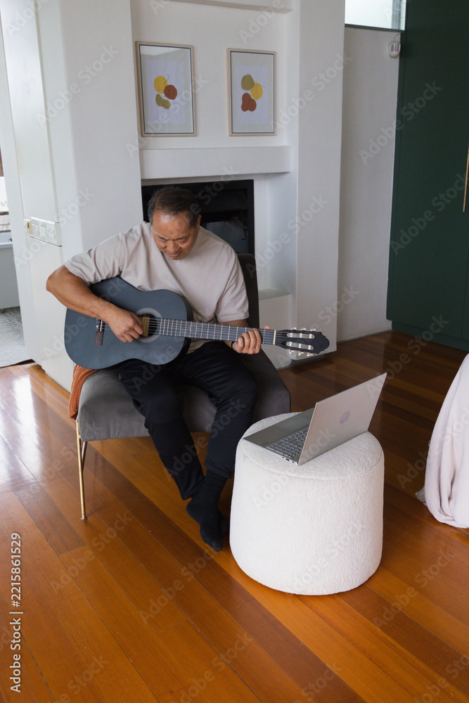 Fototapeta premium Senior Asian man playing black classical guitar in living room while referencing silver laptop