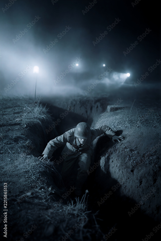 Fototapeta premium Soldier crawling out of a muddy trench at night. Vertical photo of a historical military battle with bright searchlights in thick fog. World War combat concept