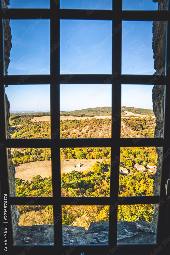 Fototapeta premium Scenic autumn landscape view framed by a medieval castle window grate. Sunny rolling hills and golden autumn trees seen through a dark iron window frame.