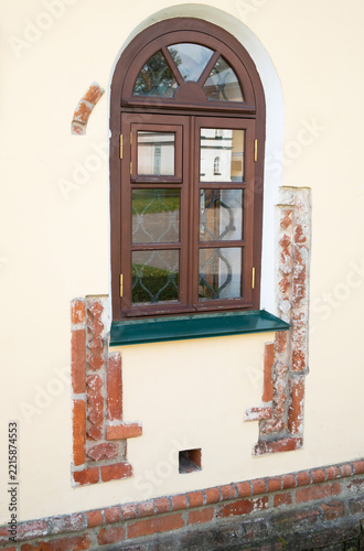 A brown window with multiple panels sits on a yellow wall. The window has a brick border and a green sill. Bricks are part of the wall design near the window.