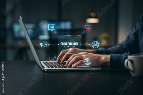 A person typing on a laptop with glowing omni channel marketing floating above the keyboard in a dimly lit office environment at night time