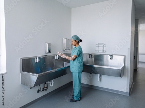 Female surgeon in medical scrubs washes her hands thoroughly at a stainless steel hospital sink before entering the operating room