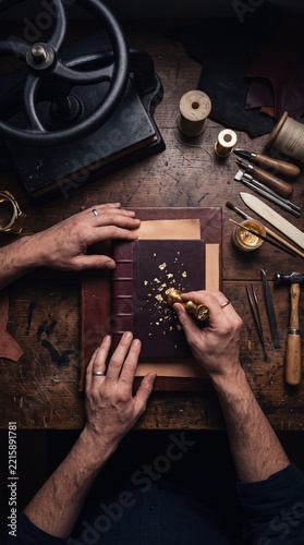 Artisan bookbinder applying gold leaf detailing onto a handmade leather journal cover at a rustic wooden workshop desk
