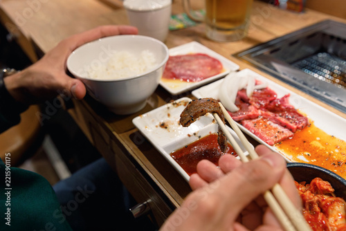 Close-up of chopsticks holding grilled beef over dipping sauce during a Japanese BBQ meal. In the background, a bowl of rice, raw sliced meat, kimchi, and a table grill.