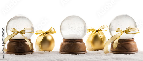 Three decorative glass snow globes with wooden bases and gold ribbon bows arranged with golden Christmas ornaments on a white snow surface