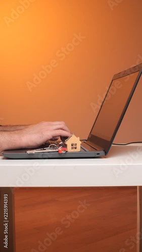 Vertical video person's hand typing on a laptop keyboard, with house keys and a house shaped keychain resting on the device, representing online home buying and property search