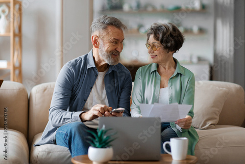 Smiling senior couple reviewing documents and smartphone together on sofa at home