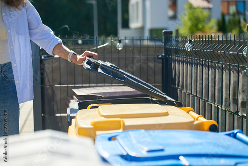 Woman opening garbage bin beside yellow and blue waste containers outside house