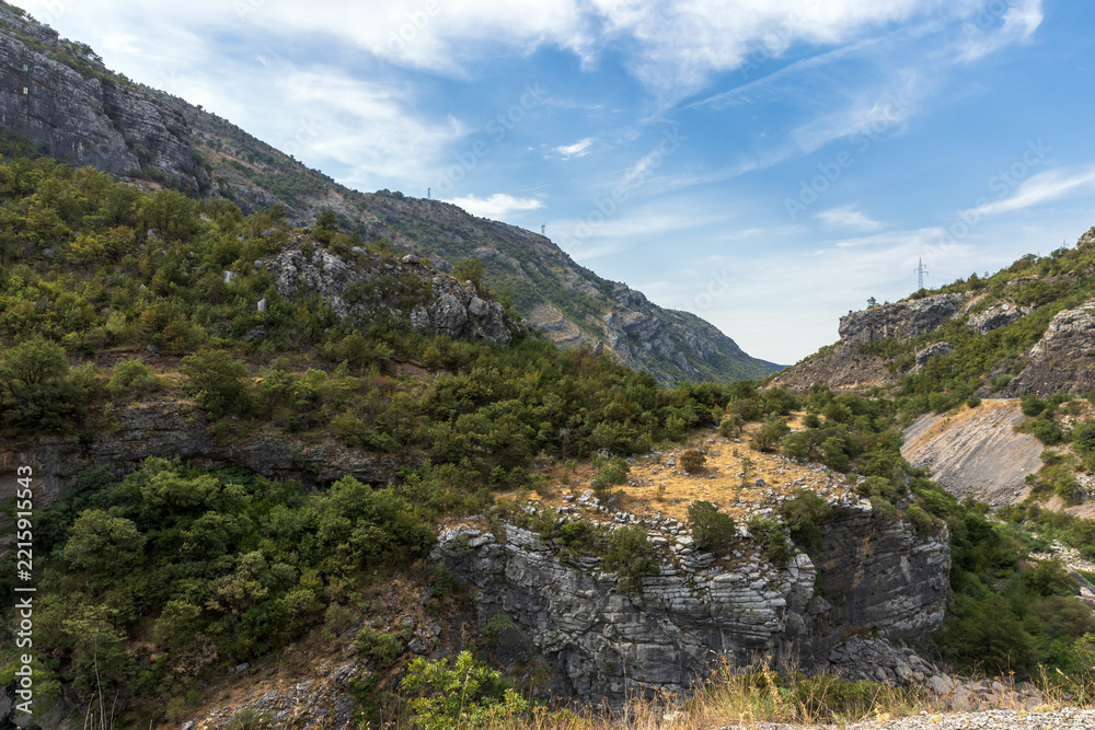 Fototapeta premium A scenic rocky canyon and valley with limestone cliffs, scrubby green hills and dry grasses beneath a clear blue sky. Rugged natural landscape suggesting hiking, geology, remote outdoors. Montenegro
