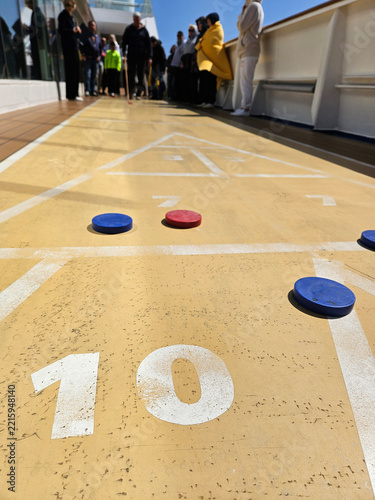 shuffleboard Sunlit deck with shuffleboard markers and discs. Players gather at distant end of marked court. Blue red discs rest near numbered section. Game setup visible on polished wooden surface