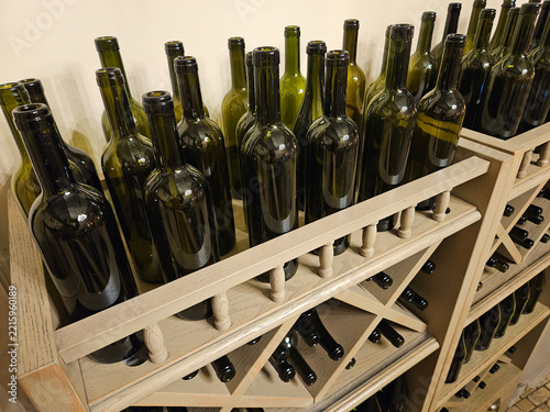 Wooden wine rack holds numerous dark glass bottles. Bottles stand upright in orderly rows. Shelves display bottles with no labels visible. Glass reflects ambient light softly