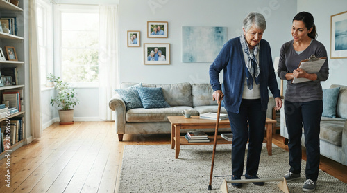 Older woman practices balance with a cane while a younger woman observes at home