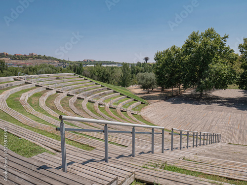View of the open-air amphitheater in Madrid's Manzanares Linear Park, featuring curved tiered seating with wooden benches and interspersed green spaces, as well as staircases with railings