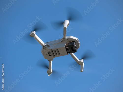 Bottom view of a small white drone with camera flying with its propellers out of focus due to speed flying through the air and a blue sky with clouds in the background