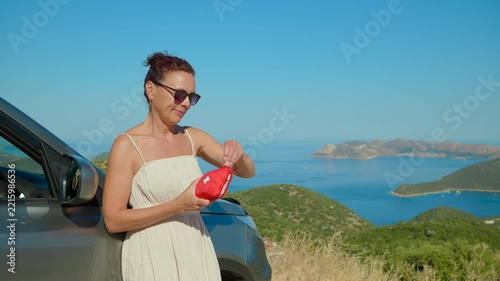 Woman traveler taking medicine from first aid kit. Woman in a summer dress standing next to her car on a scenic road trip, taking medicine from a red first aid kit against a beautiful sea and island