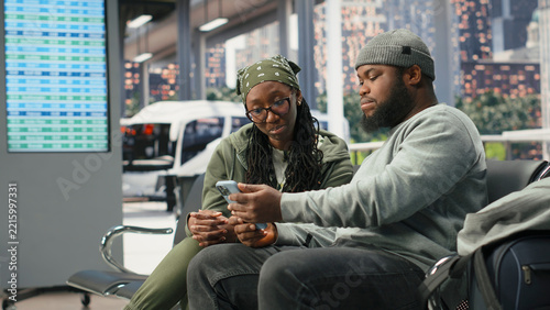 Obraz na plátně Two travelers sit together in a bus terminal and review trip plans on a smartphone, discussing their itinerary before leaving on a long-distance route