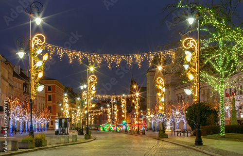 Festive Christmas illuminations decorating a historic street in Warsaw Old Town, Poland, with glowing trees and colorful holiday lights at night.