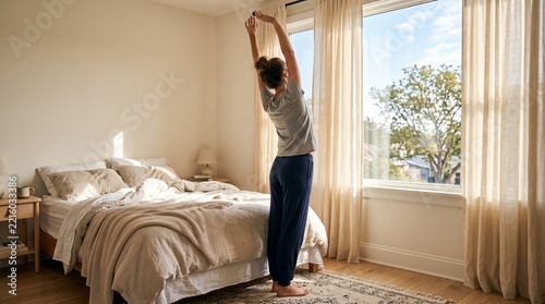 Woman waking up stretching arms by sunny bedroom window
