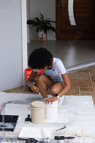 African American woman crouching on dropcloth at entry prying paint can with tool in denim overalls