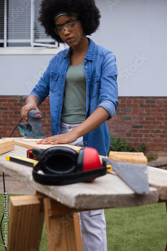 Orbital sander is smoothing wooden plank on portable workbench in backyard near brick wall