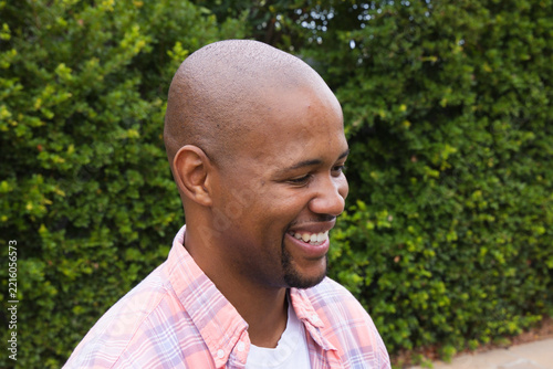 African American man smiling looking right wearing plaid shirt and white tee by trimmed green hedge
