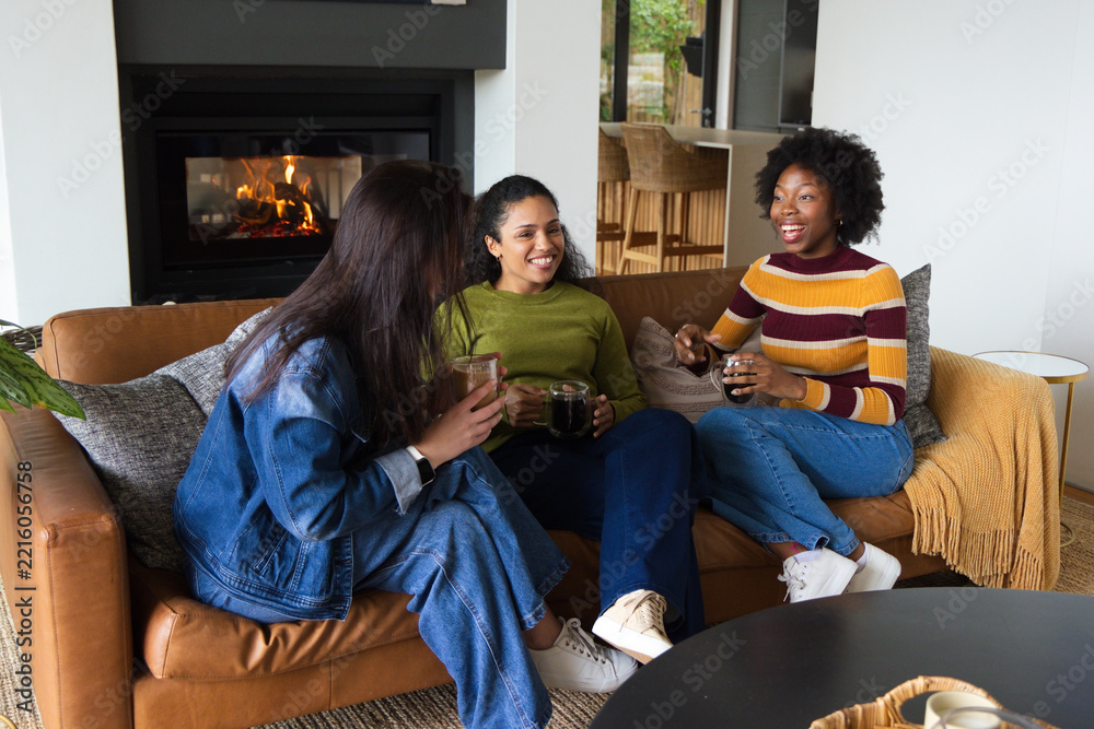 Fototapeta premium Diverse female friends sitting on leather sofa in living room, sipping from clear glass mugs