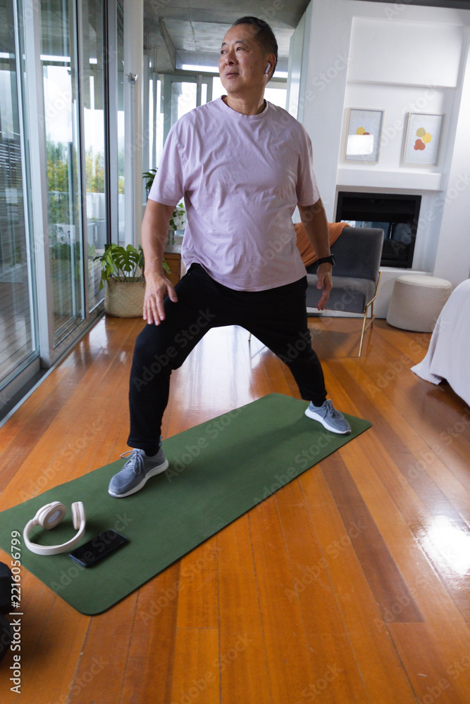 Fototapeta premium Senior Asian man stretching on green mat at home wearing pink tee black pants headphones smartwatch