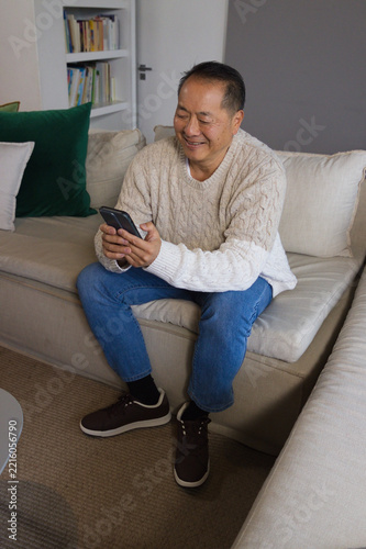 Senior Asian man sitting on beige sofa at home wearing cream sweater, jeans, holding phone smiling