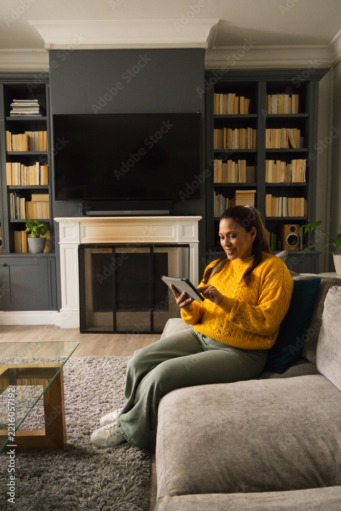 Fototapeta premium African American female sitting on sectional at home in mustard sweater using tablet, copy space