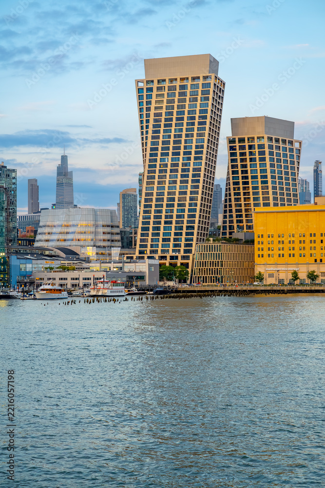 Fototapeta premium Two angled towers rise above the New York waterfront.. Brown window grids stand beside a yellow building with blue water across the vertical foreground.
