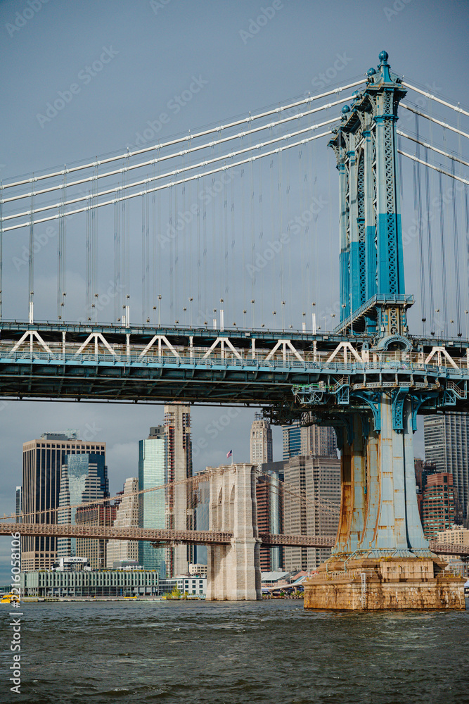Fototapeta premium A Manhattan Bridge tower frames the skyline above the river.. Steel cables cross a vertical view above a brown stone pier with the Brooklyn Bridge in the distance.