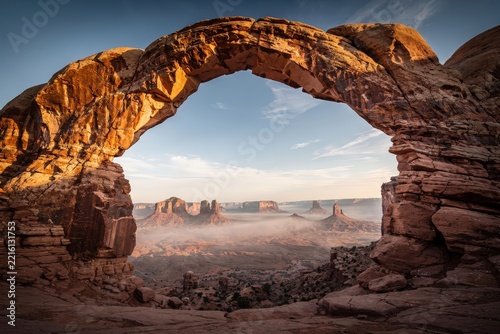 Majestic Sandstone Arch Framing Vast Desert Landscape in Monument Valley, Golden Hour Light Casting Warm Glow, Misty Horizon Enhancing Depth, Rich Earth Tones, Capturing Timeless Natural Beauty