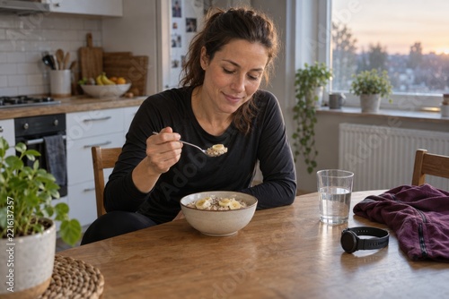 A female runner eats oatmeal with banana at her kitchen table while dressed for exercise. Balanced morning nutrition and preparation for endurance training