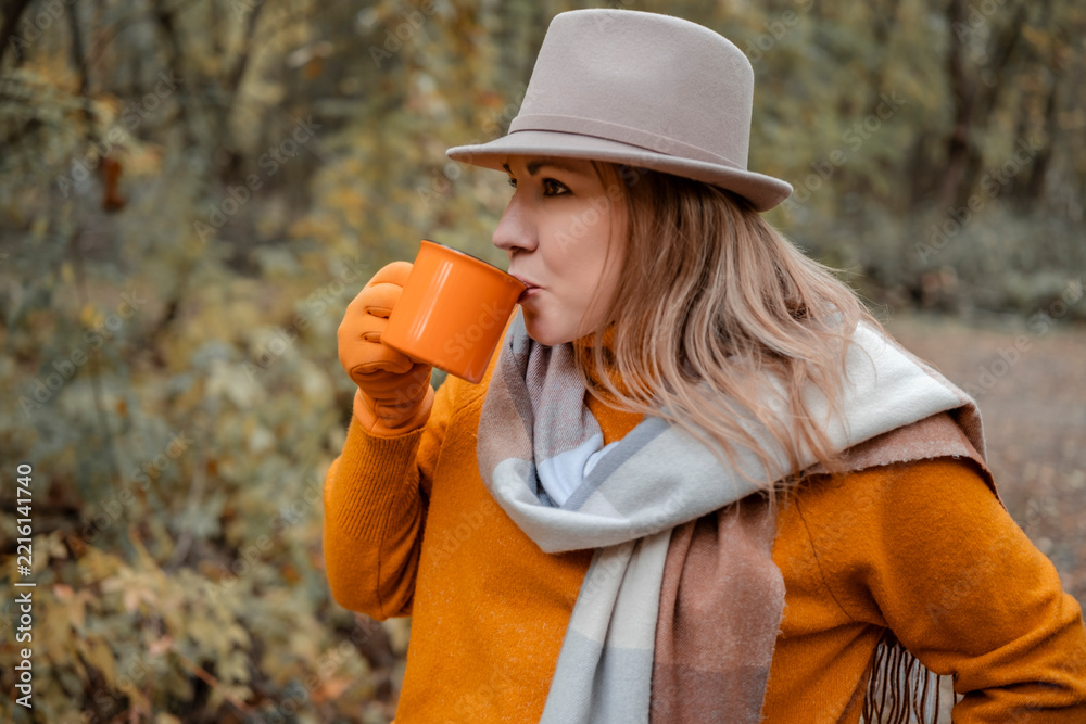 Fototapeta premium Woman drinking hot tea from mug in autumn park - cozy seasonal outdoor moment and warming beverage concept. Profile view of female in hat drinking coffee outdoors.