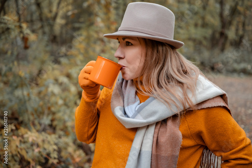 Woman drinking hot tea from mug in autumn park - cozy seasonal outdoor moment and warming beverage concept. Profile view of female in hat drinking coffee outdoors.
