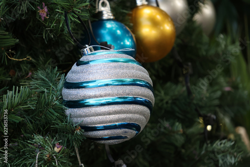 Close-up of a textured silver and metallic teal striped bauble hanging on a green Christmas tree with additional gold and blue ornaments blurred in the background
