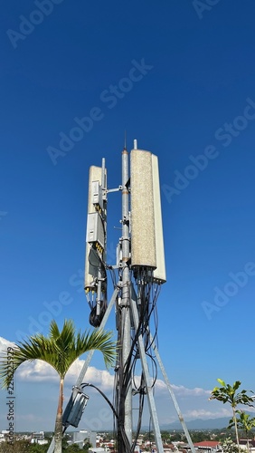 Clear blue sky with tall cellular tower structure. Multiple antennas and cables visible against the sky. Green foliage frames the bottom right corner. Ideal for telecom, infrastructure